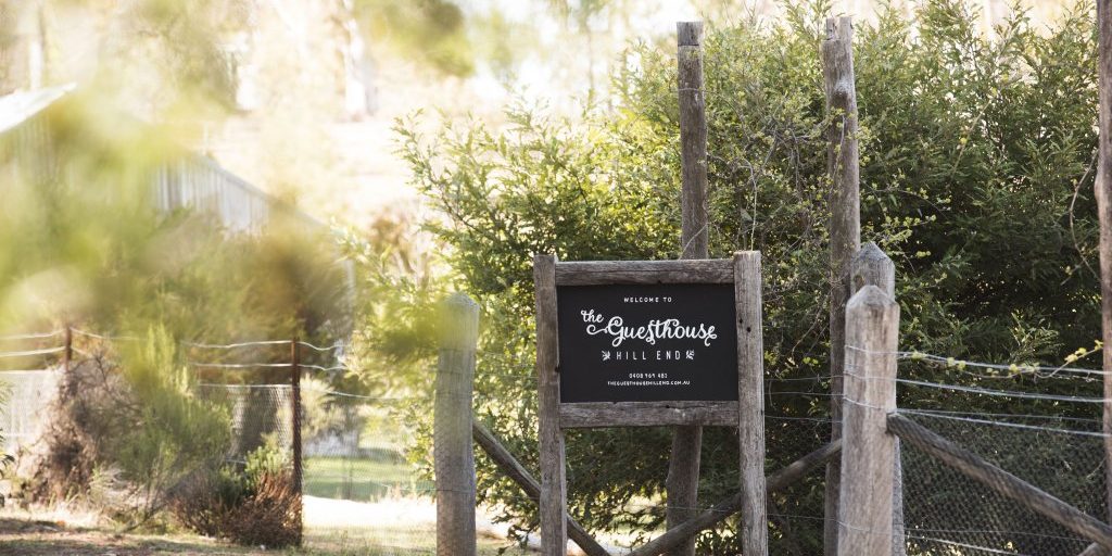 A wooden entry sign reading "The Guesthouse Hill End" stands beside a dirt pathway, surrounded by greenery and rustic fencing.