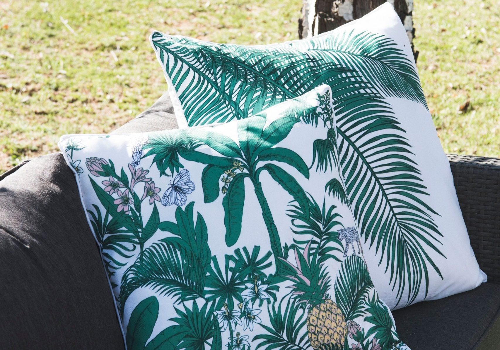 Two decorative throw pillows with green tropical leaf patterns are placed on a grey outdoor wicker sofa, situated next to a tree trunk and grassy lawn.