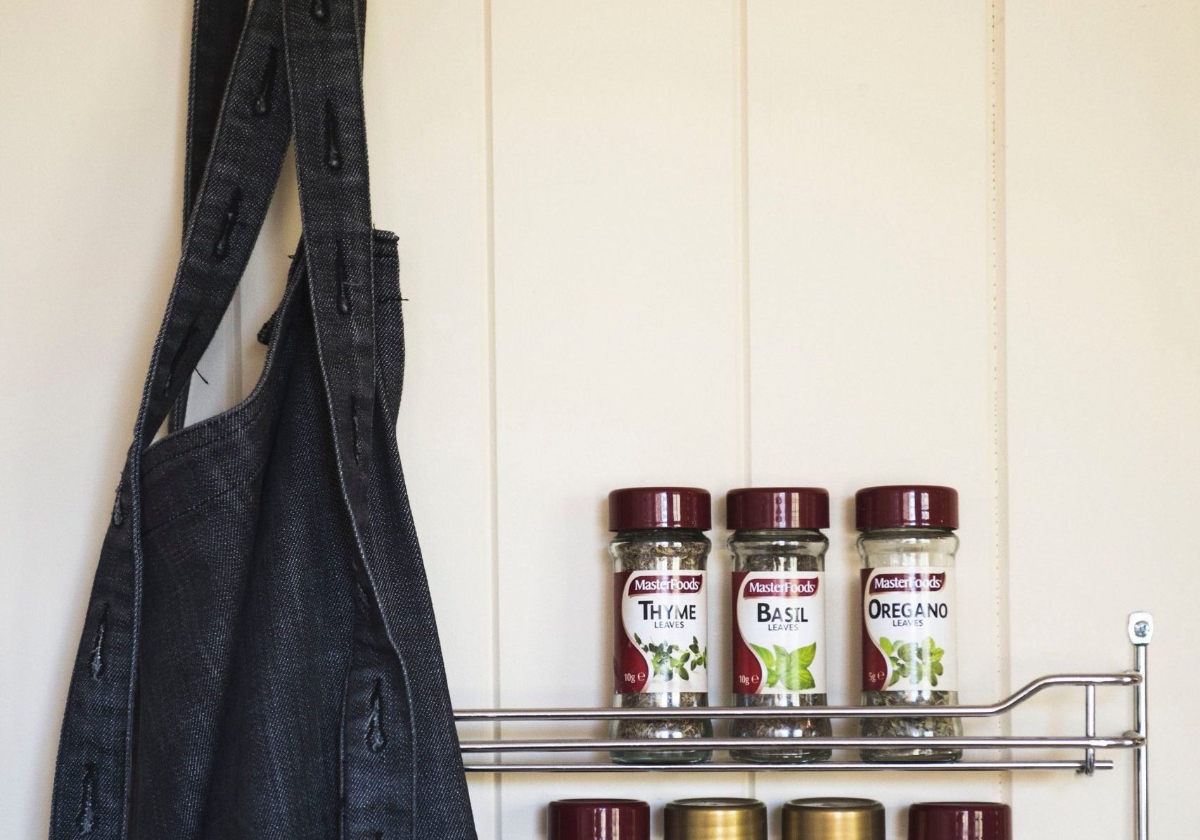 A black apron hangs next to a metal rack holding six jars of spices, including thyme, basil, oregano, garlic, Tuscan seasoning, and barbecue seasoning, on a white paneled wall.