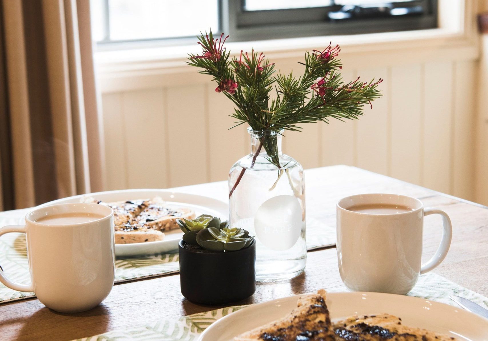 A table setting with two plates of toast topped with blueberries, two mugs, a small potted plant, and a vase with a greenery arrangement next to a window.